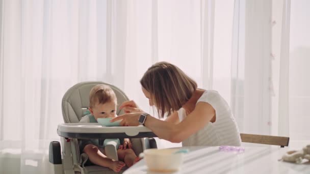 Barnet sitter vid ett litet barnbord. Mamma matar barnet med gröt. Mor ger barn mat från en sked. — Stockvideo