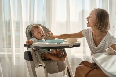 Bebek küçük bir çocuk masasında oturuyor. Annesi çocuğu yulaf lapasıyla besliyor. Anne kaşıktan bebek maması verir..