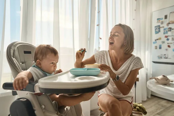 Baby sits at a childrens little table. Mum feeds the child with porridge. Mother gives baby food from a spoon. — Stock Photo, Image