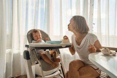 Bebek küçük bir çocuk masasında oturuyor. Annesi çocuğu yulaf lapasıyla besliyor. Anne kaşıktan bebek maması verir..