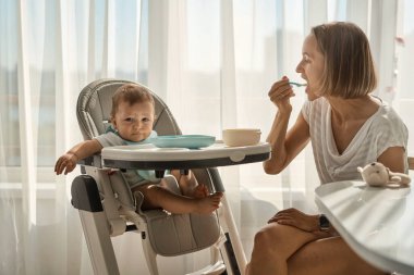 Bebek küçük bir çocuk masasında oturuyor. Annesi çocuğu yulaf lapasıyla besliyor. Anne kaşıktan bebek maması verir..