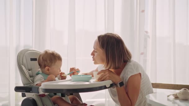 O bebê senta-se em uma pequena mesa de crianças. A mãe alimenta a criança com mingau de cereal. A mãe dá a comida de bebê de uma colher. — Vídeo de Stock