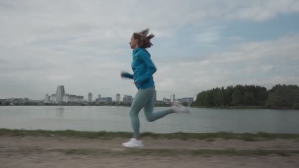 Ung idrottskvinna i sportkläder tåg i en park nära floden. Fitness kvinna jogga utomhus. Hälsosam aktiv livsstil — Stockvideo