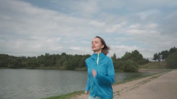 Spor giyimli genç bir bayan nehir kenarındaki bir parkta antrenman yapıyor. Spor salonundaki kadın dışarıda koşuyor. Sağlıklı aktif yaşam biçimi — Stok video