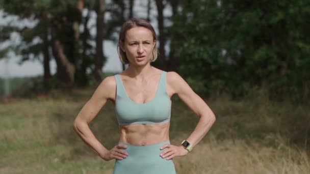 Foto de paralaje medio de mujer rubia caucásica trotando en el bosque en un día soleado. La corredora se detiene en la cima de la colina. Atleta enfocada mira al cielo — Vídeos de Stock