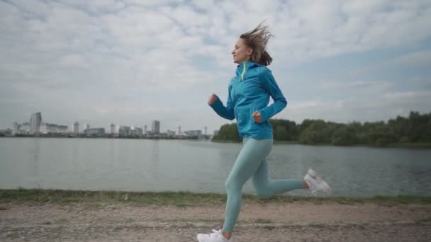 Slow motion Young sportswoman in sportswear trains in a park near the river. Fitness woman jogging outdoor. Healthy active lifestyle — Stock Video