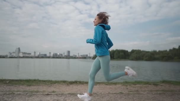 Slow motion Ung idrottskvinna i sportkläder tåg i en park nära floden. Fitness kvinna jogga utomhus. Hälsosam aktiv livsstil — Stockvideo