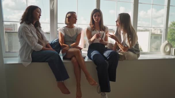 Chicas divirtiéndose en alféizar ventana. Cuatro chicas caucásicas sentadas en el alféizar de la ventana discuten algo y lough. Escuela de arte taller. — Vídeos de Stock