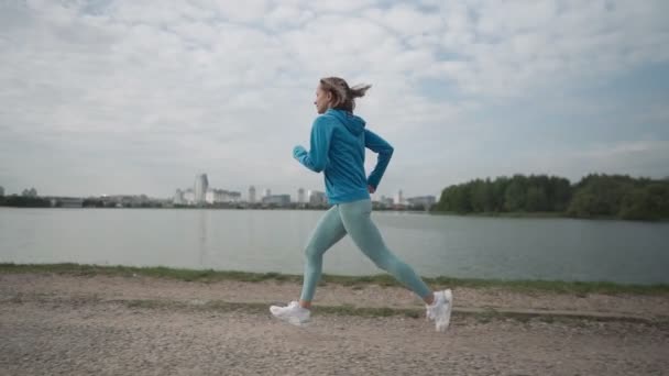 Slow motion Young sportswoman in sportswear trains in a park near the river. Fitness woman jogging outdoor. Healthy active lifestyle — Stock Video