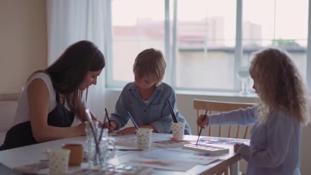 Group of kids and their teacher leaning over table with watercolor paints and painting self-made clay items. Children with teacher in art class — Stock Video