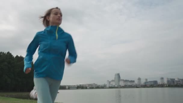 Una joven deportista en ropa deportiva entrena en un parque cerca del río. Fitness mujer corriendo al aire libre. Vida activa saludable — Vídeo de stock