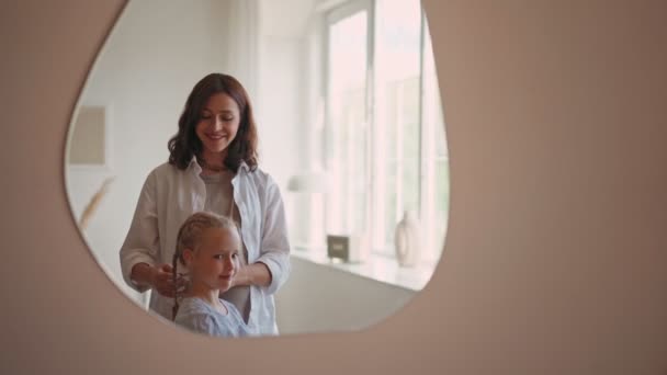 Красивая улыбающаяся девушка с кудрявыми светлыми волосами, смотрящая на свое отражение в зеркале, в то время как ее мать делает косички — стоковое видео