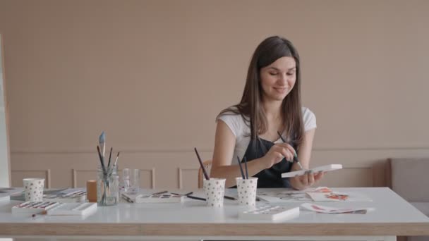 Mooie vrouwelijke kunstenares in haar atelier. Vrouwelijke tekening in album met aquarelverf en penseel. — Stockvideo