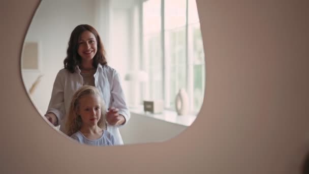 Bella ragazza sorridente con i capelli biondi ricci guardando il suo riflesso nello specchio mentre sua madre fa i capelli facendo treccia — Video Stock