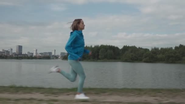 Ung idrottskvinna i sportkläder tåg i en park nära floden. Fitness kvinna jogga utomhus. Hälsosam aktiv livsstil — Stockvideo