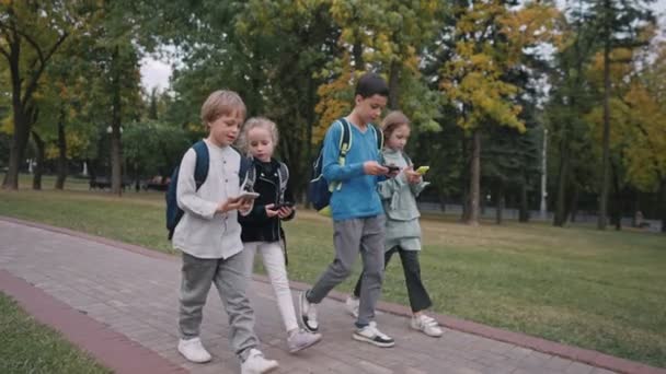 Długie ujęcie czworga uczniów z plecakami wychodzących ze szkoły i mówiących. dzieci korzystają ze smartfonów podczas chodzenia ze szkoły. — Wideo stockowe