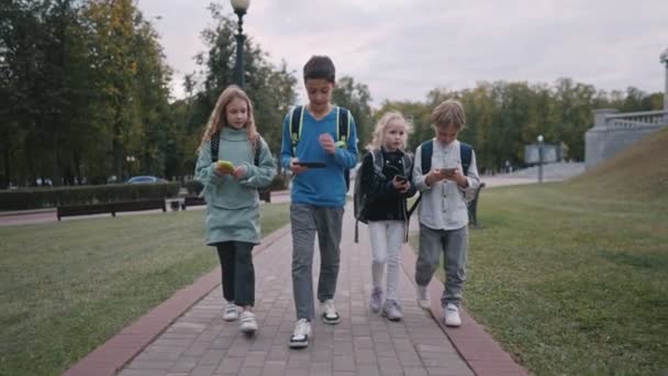 Een kleine kans van vier leerlingen met rugzakken die van school gaan en praten. kinderen gebruiken smartphones tijdens het lopen van school. — Stockvideo