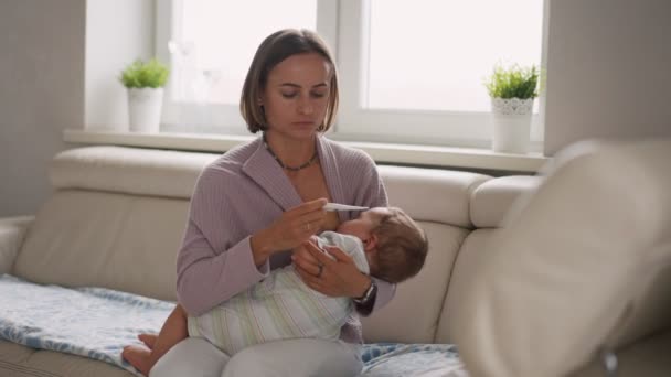 Madre joven preocupada sentada en el sofá junto a su hijo enfermo con fiebre alta. Mamá mide la temperatura usando termómetro de niño enfermo acostado debajo de una manta en casa — Vídeo de stock