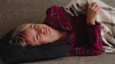 Genç bir kadın, kırmızı ekose pijamalarıyla kanepeye kıvrılmış, karnının alt kısmını görünür bir rahatsızlık içinde tutuyor. Televizyon kumandası yakında. Adet sancısı ve dinlenme dönemi kavramı. Doğal ışık, orta