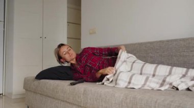 Genç bir kadın, kırmızı ekose pijamalarıyla kanepeye kıvrılmış, karnının alt kısmını görünür bir rahatsızlık içinde tutuyor. Televizyon kumandası yakında. Adet sancısı ve dinlenme dönemi kavramı. Doğal ışık, orta