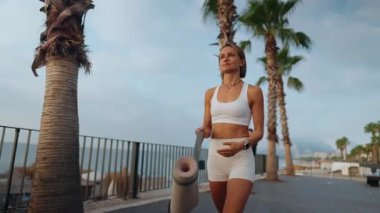 Genç Avrupalı bir kadın sabahın erken saatlerinde elinde spor minderiyle güneşli bir parkta yürüyor. Aktif giysi giyiyor ve odaklanmış ve enerjik görünüyor. Sağlıklı yaşam tarzı ve sabah rutini kavramı