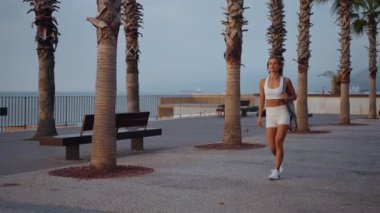 Genç Avrupalı bir kadın sabahın erken saatlerinde elinde spor minderiyle güneşli bir parkta yürüyor. Aktif giysi giyiyor ve odaklanmış ve enerjik görünüyor. Sağlıklı yaşam tarzı ve sabah rutini kavramı