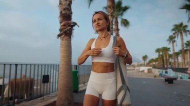 Genç Avrupalı bir kadın sabahın erken saatlerinde elinde spor minderiyle güneşli bir parkta yürüyor. Aktif giysi giyiyor ve odaklanmış ve enerjik görünüyor. Sağlıklı yaşam tarzı ve sabah rutini kavramı