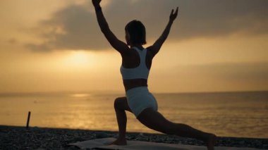 Genç bir kadın gün doğumunda plajda yoga yapıyor, yukarı bakan köpek pozu veriyor. Beyaz etek giyiyor ve dikkatle ve zarafetle hareket ediyor. Sabah ritüeli, güç ve iç güdü kavramı