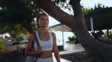 Genç Avrupalı bir kadın sabahın erken saatlerinde elinde spor minderiyle güneşli bir parkta yürüyor. Aktif giysi giyiyor ve odaklanmış ve enerjik görünüyor. Sağlıklı yaşam tarzı ve sabah rutini kavramı