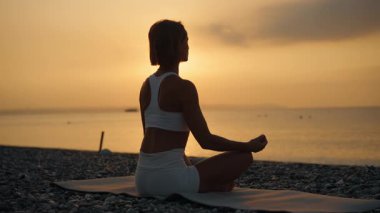 Genç bir kadın, huzurlu bir sabah güneşi doğarken deniz kenarındaki bir minderde yoga yapıyor. Suya bakan meditasyon poziyonunda oturuyor. Farkındalık, iç denge ve doğayla bağlantı kavramı