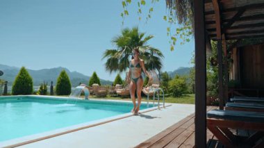 Güneşli bir yaz gününde, lüks bir açık havuzun yanında bikinili atletik genç bir kadın yürüyor. Etrafı yemyeşil ve dağ manzaralarıyla çevrili, lüks bir yerde huzurlu bir tatilin tadını çıkarıyor.