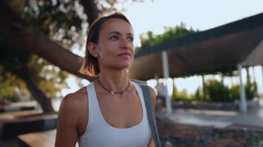 Genç Avrupalı bir kadın sabahın erken saatlerinde elinde spor minderiyle güneşli bir parkta yürüyor. Aktif giysi giyiyor ve odaklanmış ve enerjik görünüyor. Sağlıklı yaşam tarzı ve sabah rutini kavramı