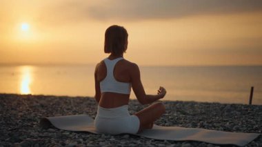 Genç bir kadın, huzurlu bir sabah güneşi doğarken deniz kenarındaki bir minderde yoga yapıyor. Suya bakan meditasyon poziyonunda oturuyor. Farkındalık, iç denge ve doğayla bağlantı kavramı
