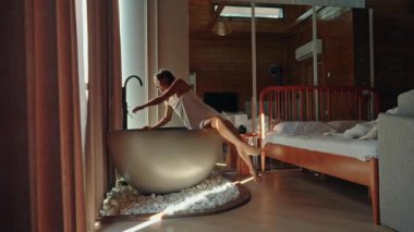 Havluya sarılmış genç bir kadın sıcacık bir ahşap evde sıcak bir köpük banyosu hazırlıyor. Sabah rahatlama ve kişisel bakım ayini. Doğal ışık odayı dolduruyor ve huzurlu bir atmosfer yaratıyor.