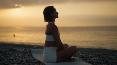 Genç bir kadın, huzurlu bir sabah güneşi doğarken deniz kenarındaki bir minderde yoga yapıyor. Suya bakan meditasyon poziyonunda oturuyor. Farkındalık, iç denge ve doğayla bağlantı kavramı