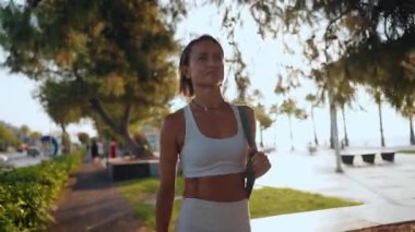 Genç Avrupalı bir kadın sabahın erken saatlerinde elinde spor minderiyle güneşli bir parkta yürüyor. Aktif giysi giyiyor ve odaklanmış ve enerjik görünüyor. Sağlıklı yaşam tarzı ve sabah rutini kavramı