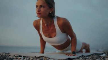 Genç bir kadın gün doğumunda plajda yoga yapıyor, yukarı bakan köpek pozu veriyor. Beyaz etek giyiyor ve dikkatle ve zarafetle hareket ediyor. Sabah ritüeli, güç ve iç güdü kavramı