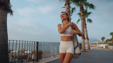 Genç Avrupalı bir kadın sabahın erken saatlerinde elinde spor minderiyle güneşli bir parkta yürüyor. Aktif giysi giyiyor ve odaklanmış ve enerjik görünüyor. Sağlıklı yaşam tarzı ve sabah rutini kavramı