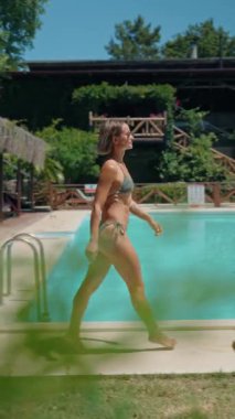 Güneşli bir yaz gününde, lüks bir açık havuzun yanında bikinili atletik genç bir kadın yürüyor. Etrafı yemyeşil ve dağ manzaralarıyla çevrili, lüks bir yerde huzurlu bir tatilin tadını çıkarıyor.