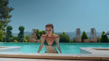 Güneşli bir yaz gününde bikinili genç bir kadın havuzdan çıkıyor. Etrafı yeşillik ve dağlarla çevrili, huzurlu tatil atmosferlerinden hoşlanıyor. Sağlık kavramı, aktif yaşam tarzı ve açık hava.