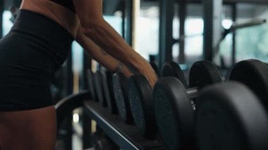 Modern bir spor salonunun rafından dambıl seçen atletik bir kadının yakın çekimi. Görüntü, dayanıklılık eğitiminden, odaklanmadan, niyetten ve disiplinden önceki hazırlayıcı anı yakalar. Yüksek