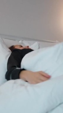 Sabahları büyük bir otel yatağında beyaz bir yatağa sarılmış ve yumuşak yastıkların üzerinde dinlenen bir kadın. Sıcak ışıklandırma ve asgari dekor ile modern iç mekan sakin ve rahat bir atmosfer yaratır. O