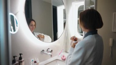 Kadın sabah bakım rutini sırasında bir damlatıcıyla kafa derisi tedavisiyle aynanın karşısına geçiyor. Modern bir banyoda bornoz giyerek dikkatlice sıvı bir serum dağıtır.