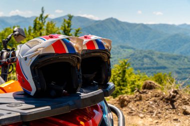 Arka planda orman tepeleri ve manzaralı vadi olan dağlarda duraklarken dört tekerli bisiklete yerleştirilmiş iki kask. Macera seyahati konsepti, off-road keşfi ve açık hava motor yolculuğu için güvenlik teçhizatı.