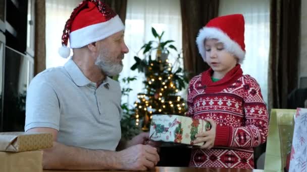 Boldog medve idősebb férfi piros karácsonyi kalapban az unokájával és ajándékokkal, ünnepli az új évet — Stock videók