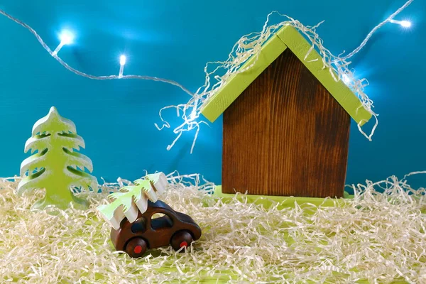 The wooden model of car with fir tree on is on the background of wooden model of house among winter landscape. The concept of Christmas, New Year, holiday, winter vacation, travel.