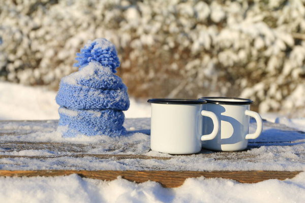 A two white iron mugs of tea next to blue knitted hat and scarf stand on wooden table on the background of winter landscape.  