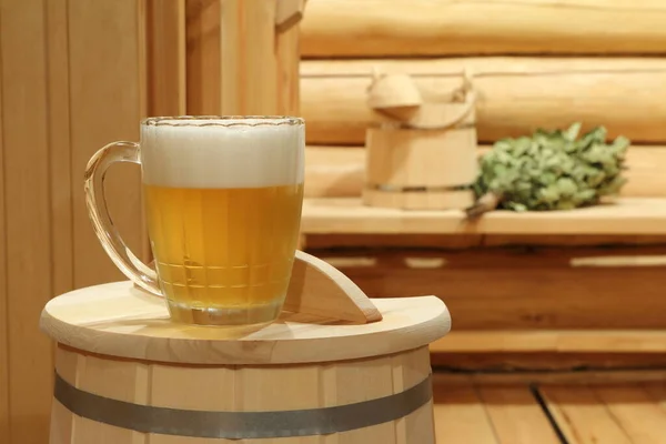 木製のベンチにはサウナのアクセサリーを背景に ライトビールのグラスマグカップだけが木製のバケツに乗っています — ストック写真