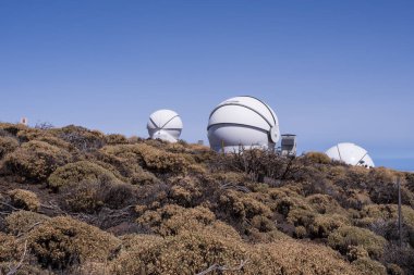 Tenerife volkanik yamaçlarındaki modern teleskop kubbeleri bulutların üzerindedir. Kuru çalılar, mavi gökyüzü ve Kanarya Adaları 'ndaki Teide Ulusal Parkı yakınlarındaki bilim araştırma tesisi..