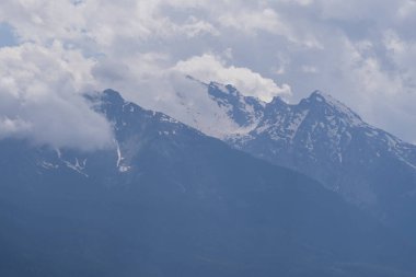 Karla kaplı zirveler, çarpıcı bulutlarla dolu yemyeşil orman yamaçlarının üzerinde duruyor. Seyahat, yürüyüş, irtifa ve manzara temaları için ideal barışçıl bir dağ manzarası..